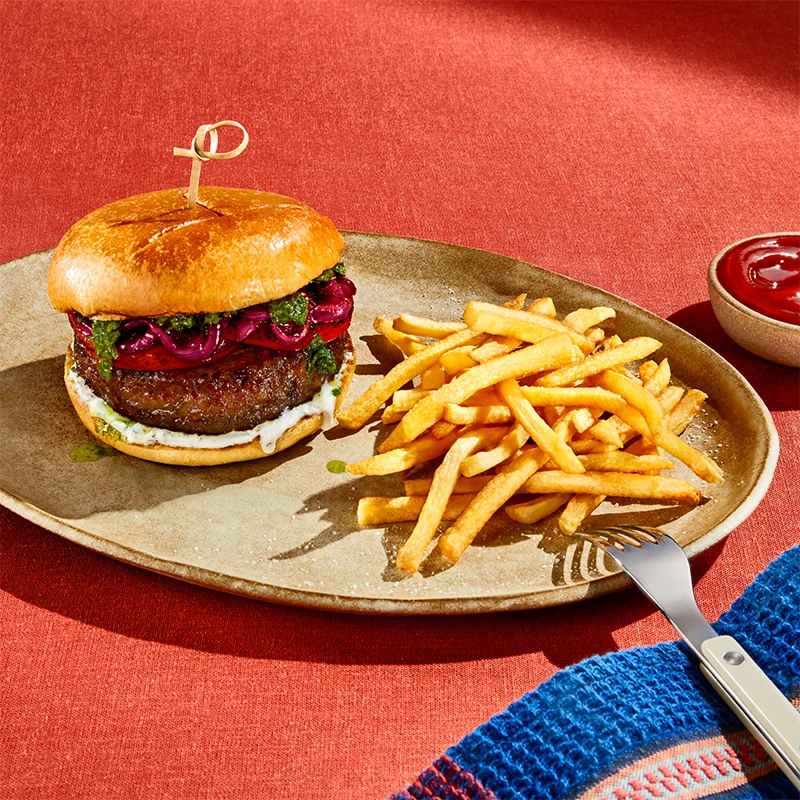 A juicy, thick Impossible™ Indulgent Burger patty with goat cheese aioli, charred tomato slices, red onion jam, and green pistou sausage, all on a toasted brioche bun, served on a plate with a side of crispy fries and ketchup. 
