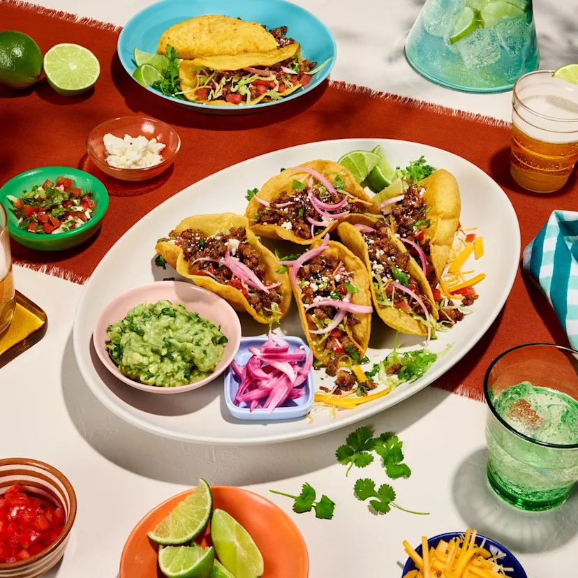 A platter of Impossible Crispy Tacos dressed with pico de gallo, lettuce, and cotija cheese, served alongside fresh lime wedges and guacamole. 