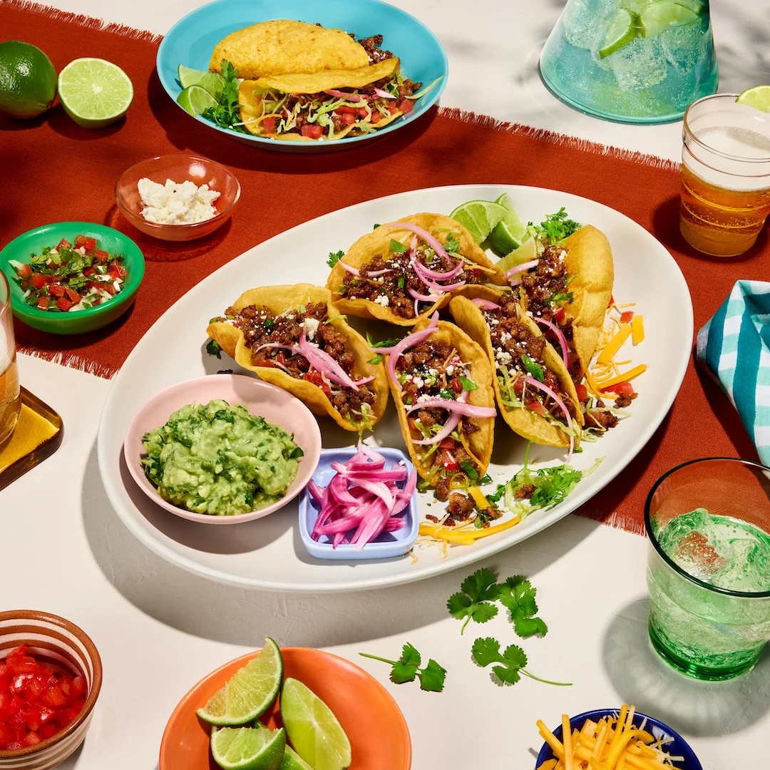 A platter of Impossible Crispy Tacos dressed with pico de gallo, lettuce, and cotija cheese, served alongside fresh lime wedges and guacamole.