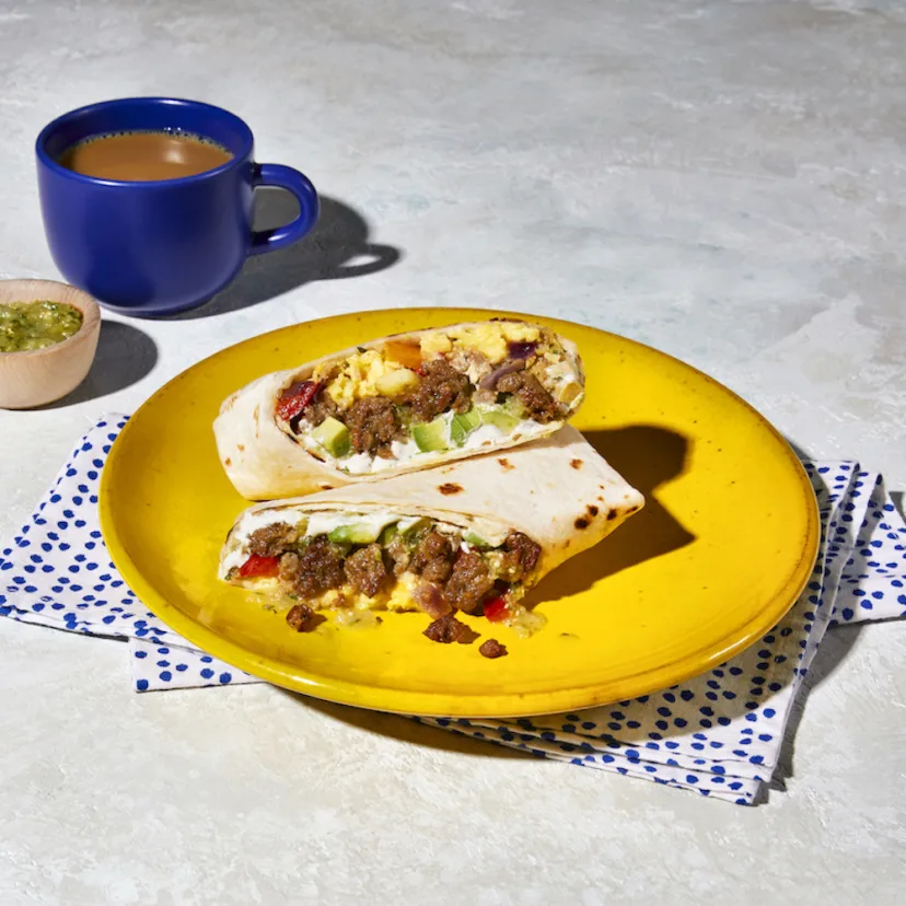 A cut open Impossible™ Sausage Breakfast Burrito with scrambled egg, Impossible Sausage, avocado, salsa, and cilantro lime crema, on a yellow plate with a napkin and a cup of coffee and side of tomatillo salsa. 