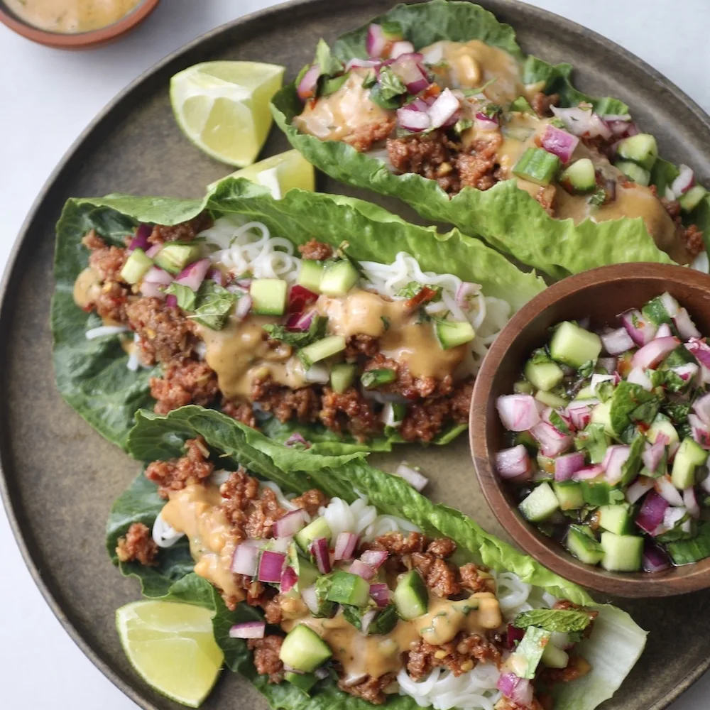 Three Impossible Beef Lettuce Cups on a plate with lime wedges, filled with rice noodles, spiced plant-based beef, peanut sauce, and a fresh cucumber onion salad. 