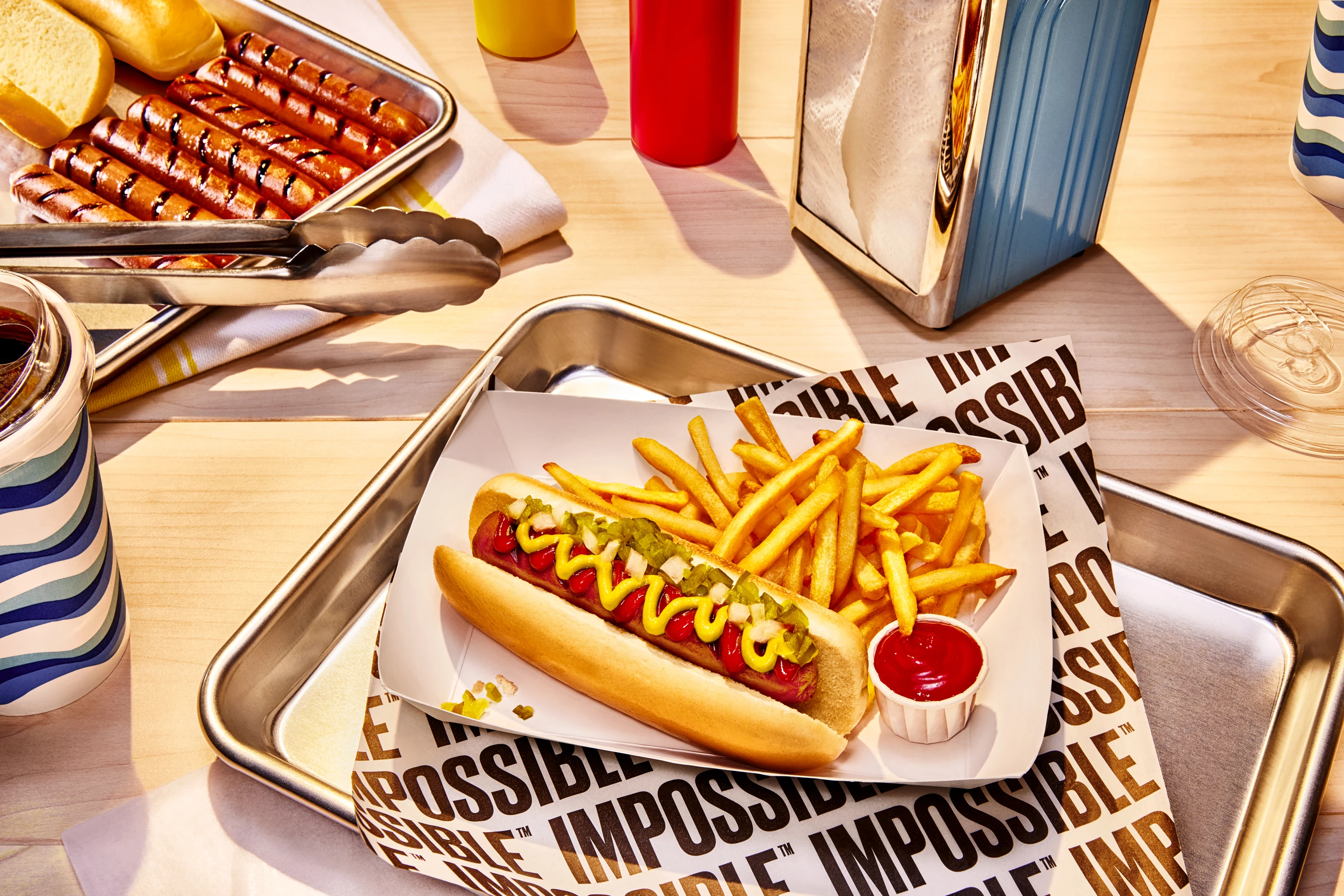 hotdog with mustard, onions, and relish on it. Sitting with french fries and side of ketchup on a paper bowl on top of impossible foods logos paper in a metal tray on a diner table. 