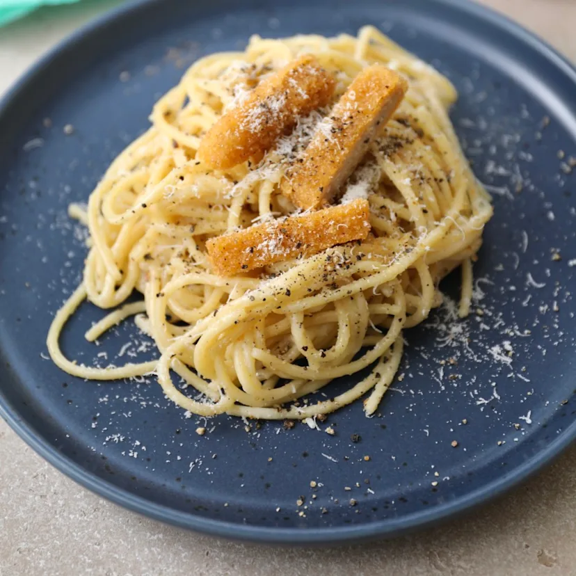 A plate of Impossible™ Chicken Nuggets Cacio e Pepe, with spaghetti, black pepper and grated cheese, topped with slices of crispy Impossible Chicken Nuggets. 