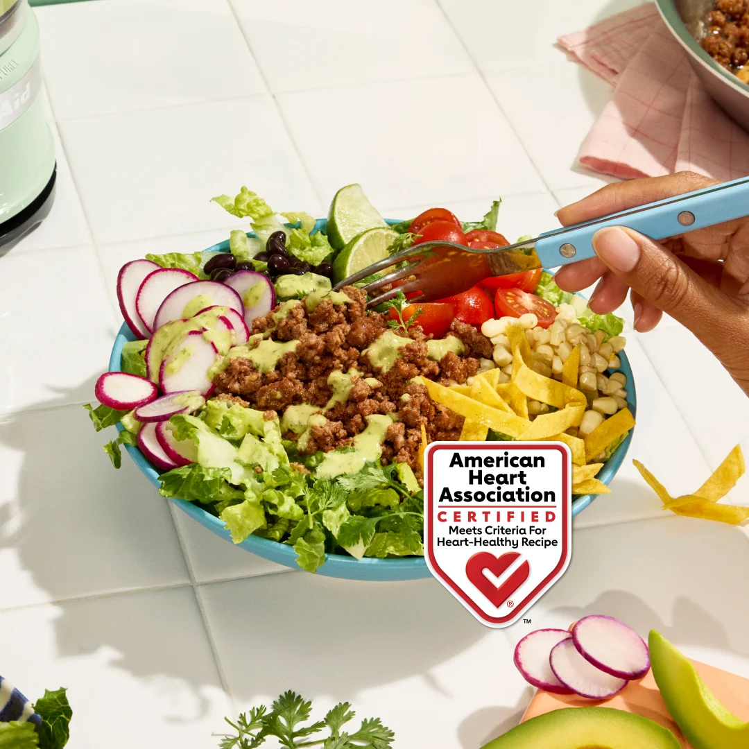 A large bowl of taco salad with taco spiced Impossible Lite Ground Beef Meat From Plants, sliced red radishes, crisp romaine, cherry tomatoes, black beans, white corn, tortilla strips, dressing with an avocado lime dressing.