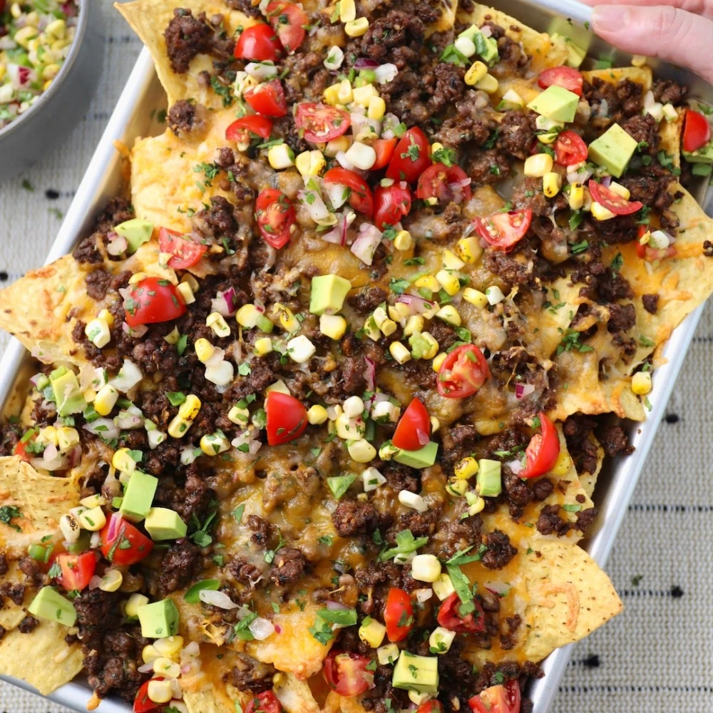 Tortilla chips on a sheet pan covered in chorizo spiced Impossible™ Beef with corn salsa, avocado and tomatoes, all held together with melted cheese. 