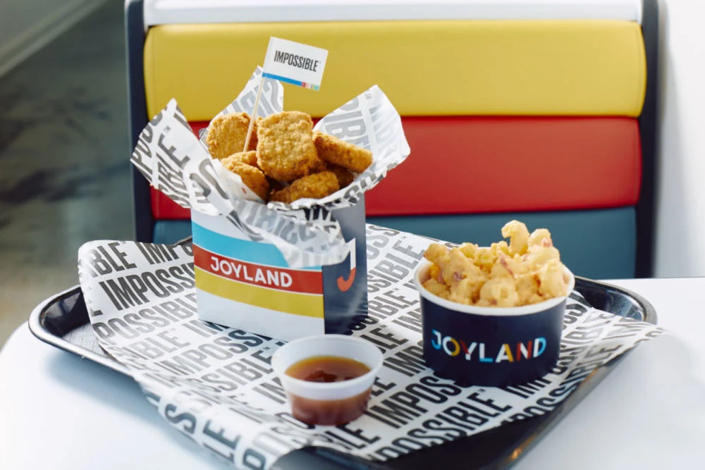 Impossible™ Chicken Nuggets Made From Plants, Peach Sweet & Sour Dipping Sauce, and Pimento Mac & Cheese in colorful Joyland Restaurant packaging