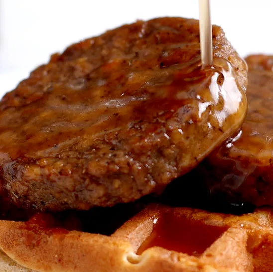 A crispy waffle on a plate topped with two Impossible Sausage Patties, drizzled with buttered maple syrup. 