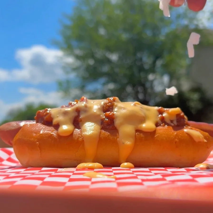 A cheese sauce-dripping chili cheese hot dog on a bun, featuring Impossible Beef Chili and Impossible Beef Hot Dogs.