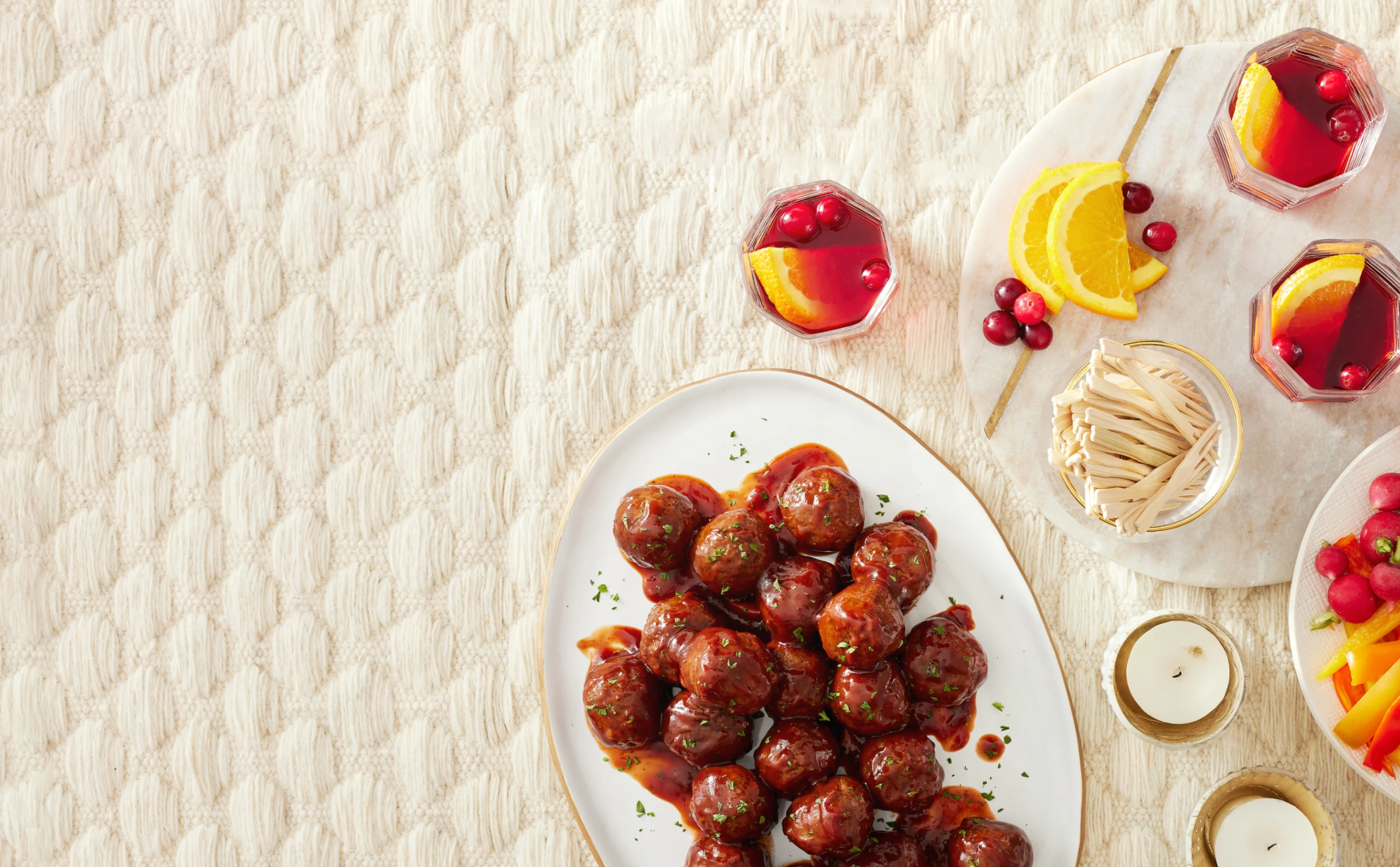 meatballs on a cream colored quilt background. cranberry and orange cocktails around the platter of meatballs