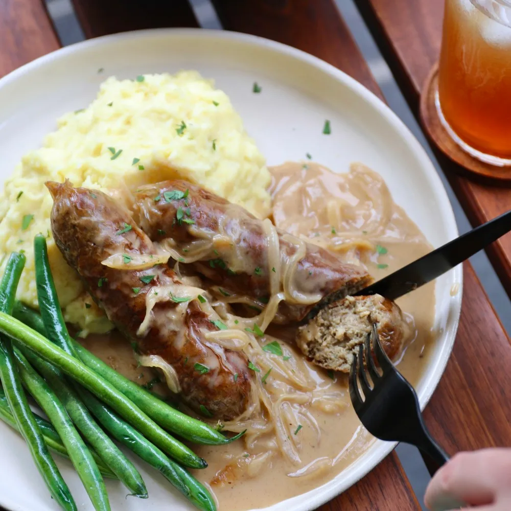 Two Impossible™ Sausage Links with skillet onion gravy on top of mashed potatoes and green beans, and garnished with chopped parsley. 