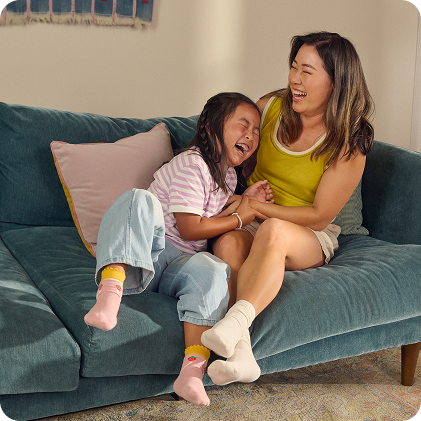 A woman and a young girl sit together on a couch in a sunlit living room. They are laughing and holding hands. 
