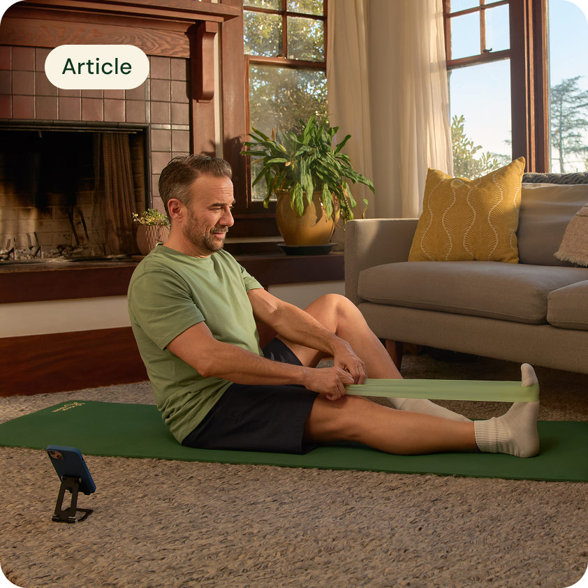 A man is sitting on a green yoga mat in his sunlit living room and is stretching with an exercise band.