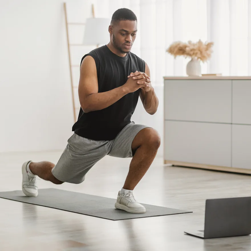 Person doing leg mobility exercises on exercise mat at home, following workout on laptop, with water bottle and jump rope nearby