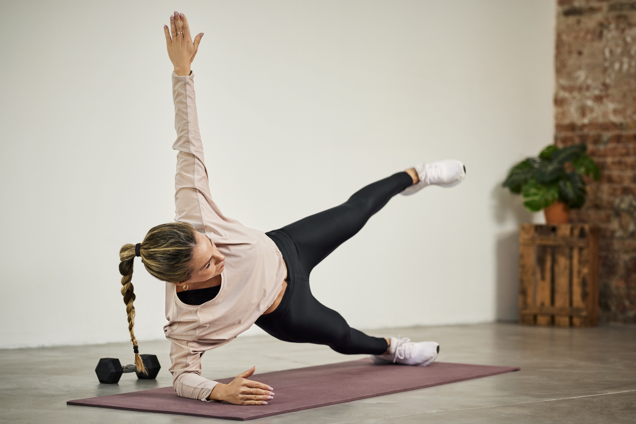 Woman in pink top and black leggings performing hip exercises for runners on purple yoga mat, with dumbbell nearby