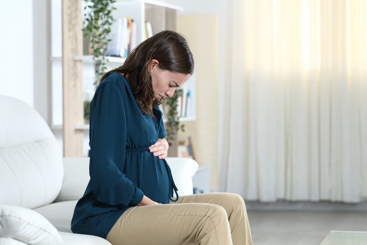 Person in teal dress sitting on white couch experiencing abdominal discomfort due to lightning crotch during pregnancy. 