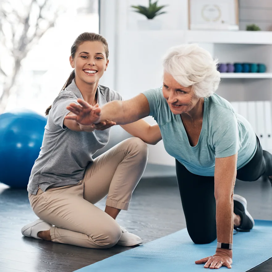 Physical therapist guides senior woman during balance exercise on yoga mat for physical therapy after back surgery.
