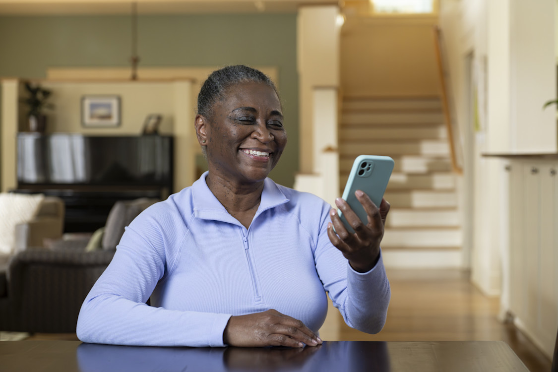 Image of a woman with short gray hair seated at a table in her home with a living room behind her. She's smiling at the phone she holds in her left hand.