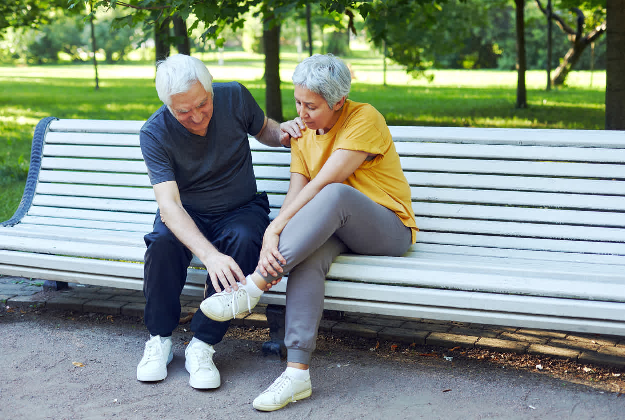 elderly-man-and-woman-sitting-in-bench-at-park-touching-ankle
