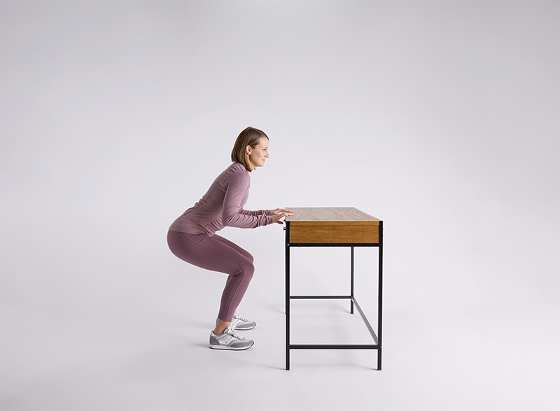 Person in mauve athletic wear practicing knee replacement exercises using a wooden desk for support against white background