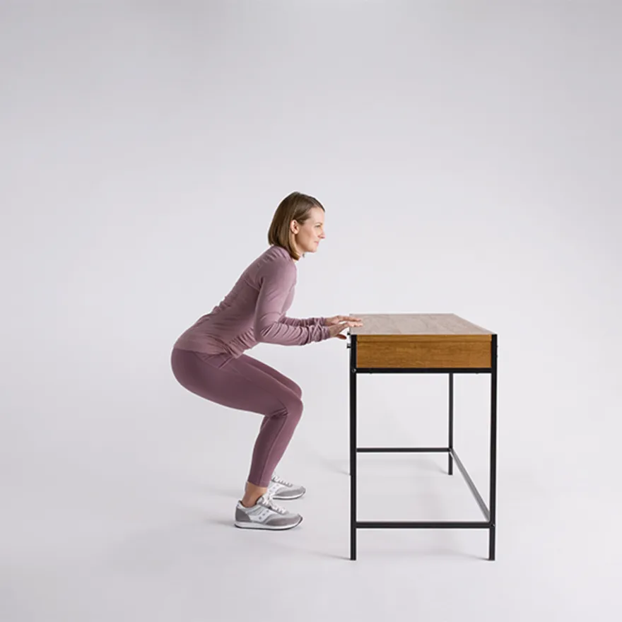 Person in mauve athletic wear practicing knee replacement exercises using a wooden desk for support against white background