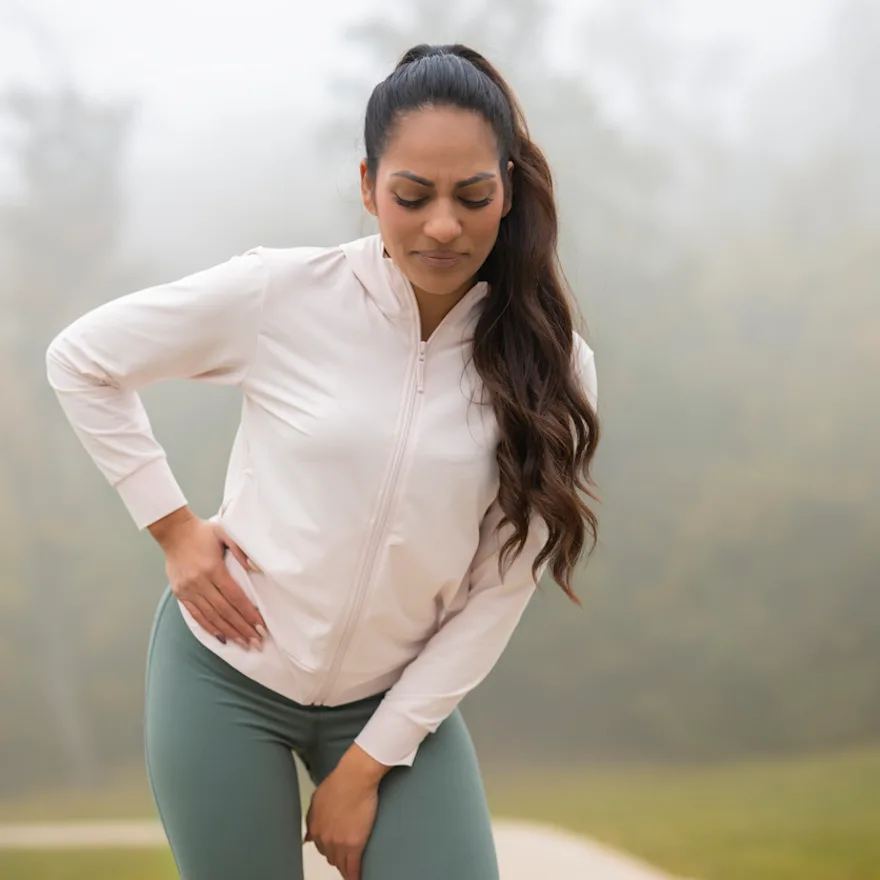Woman in white zip-up jacket and green leggings experiencing pain in front of hip.