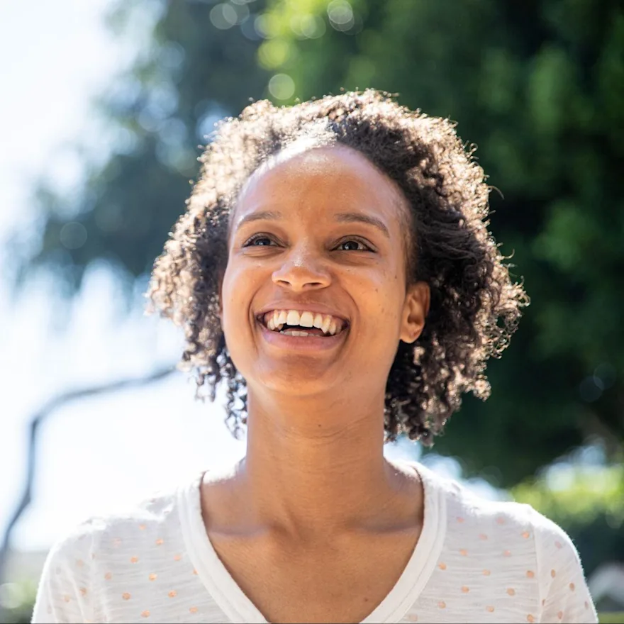 Person with natural curly hair laughing joyfully because she knows her worth, she's outdoors on sunny day, wearing white polka dot top