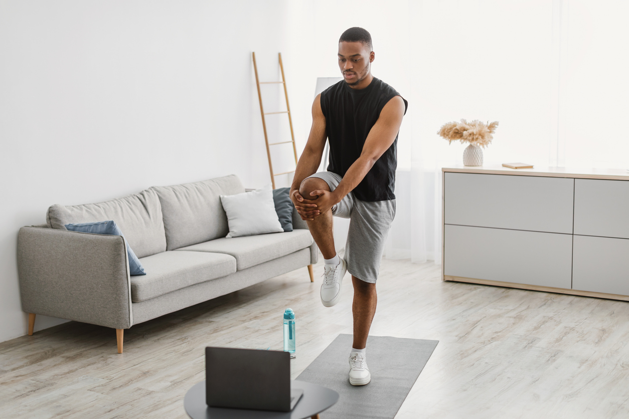 Person practicing MCL exercises, stretching leg while watching laptop in modern living room with gray sofa and minimalist decor