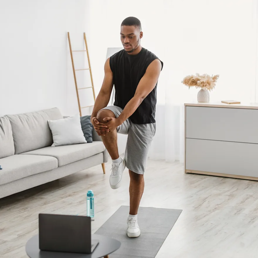 Person practicing MCL exercises, stretching leg while following workout on laptop in modern living room with gray sofa