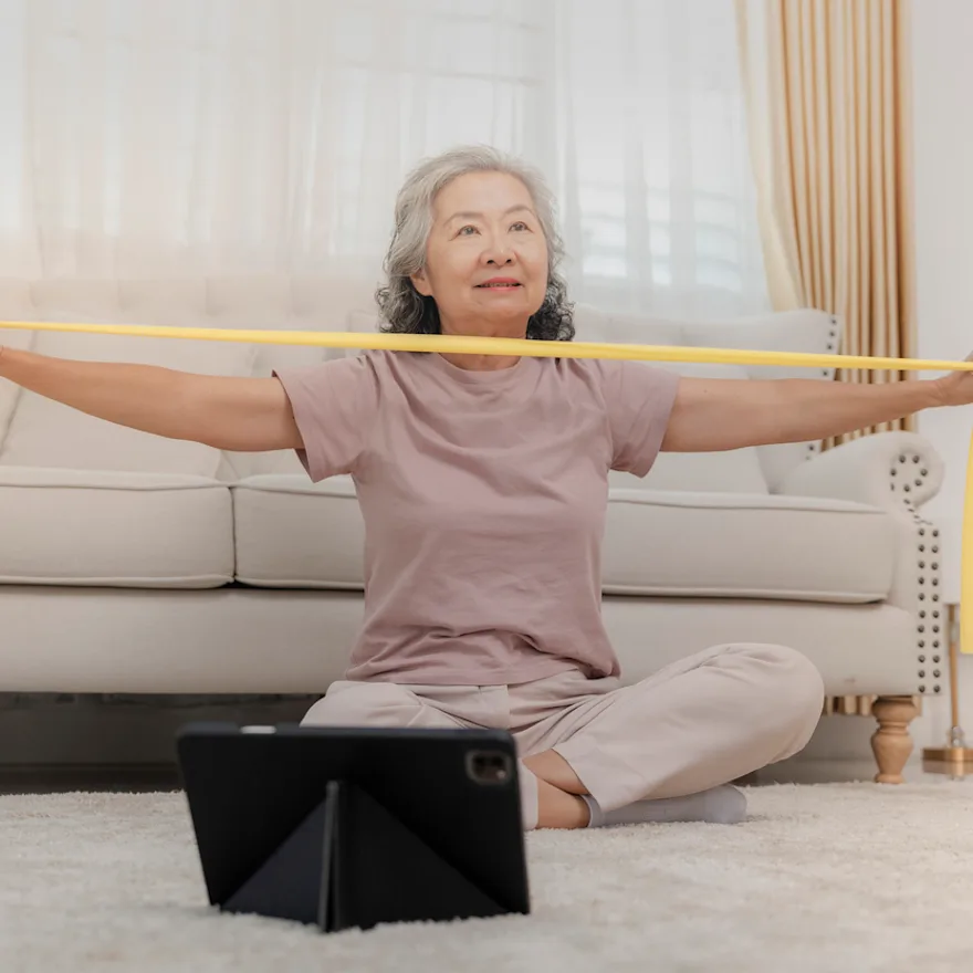 Senior woman exercising at home with yellow resistance band while seated on floor near white couch, wearing beige workout clothes