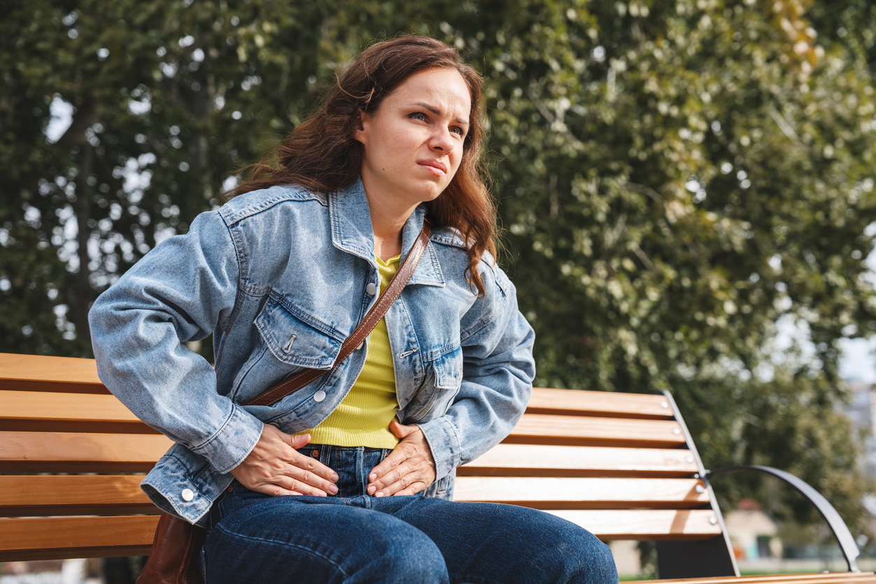 Person in denim jacket and yellow top sitting on wooden bench outdoors, experiencing pelvic pain when walking. 
