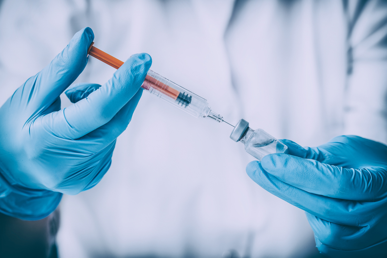 Gloved hands preparing joint injection, drawing orange liquid from vial into syringe against blurred white background
