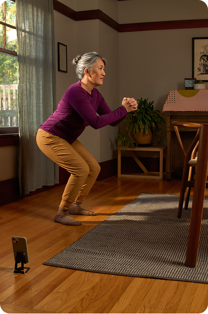 A middle-aged woman is doing squats in her living room while a closeup of the Hinge Health app is showing the same exercise.