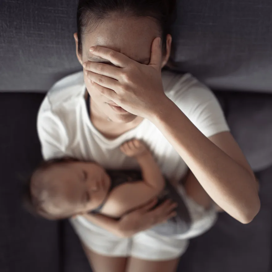 Person in white clothing holding sleeping baby while covering face with hand due to postpartum headaches.