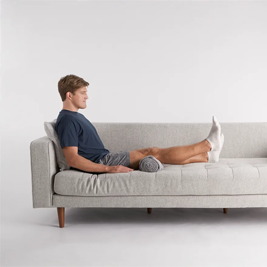 Person reclining on light gray modern sofa with rolled blanket under knees, practicing hip replacement exercises.