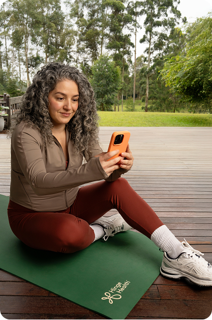 A woman is sitting on a green yoga mat outside in a park and a closeup of the Hinge Health app is showing a cat cow exercise.