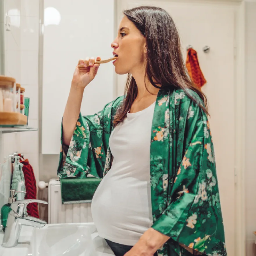 Pregnant woman in floral green robe brushing teeth in bathroom, wearing white tank top, standing at sink with mirror visible