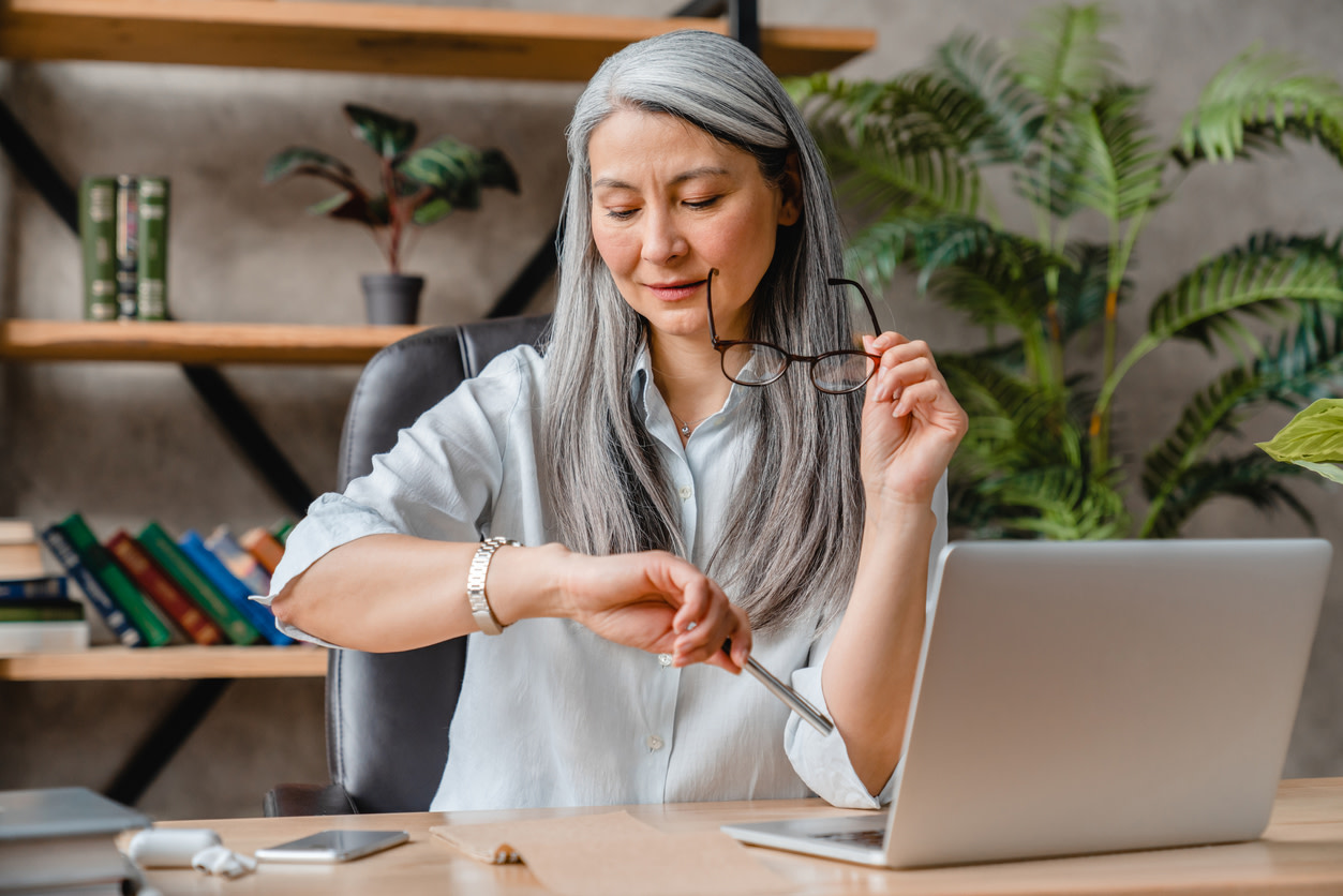 Image of a middle-aged woman woman sitting at a desk in front of her computer with a bookshelf and a green plant visible behind her. She's holding a pair of glasses in one hand and looking to check the time on the watch on her wrist.
