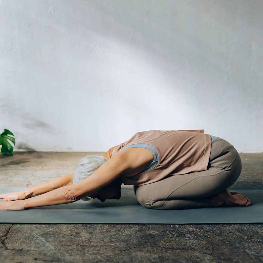 Person performing exercises for constipation on gray yoga mat against white wall with monstera plant in corner, minimalist home yoga setting