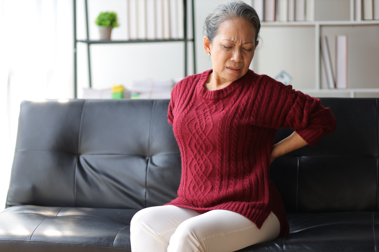 Senior woman in red sweater sitting on black couch, grimacing while holding her lower back due to thoracic spondylosis.