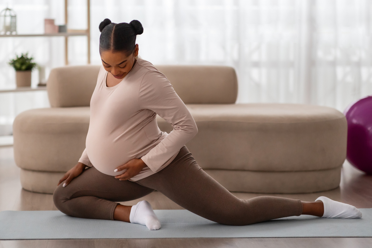 Pregnant woman in pink top and brown leggings stretches on yoga mat at home, performing gentle prenatal exercise