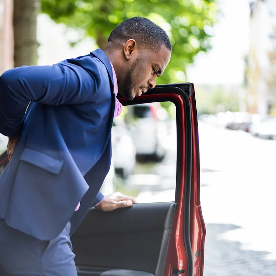 Professional man in navy blue suit entering a red car on a sunny day, with trees and buildings visible in the background