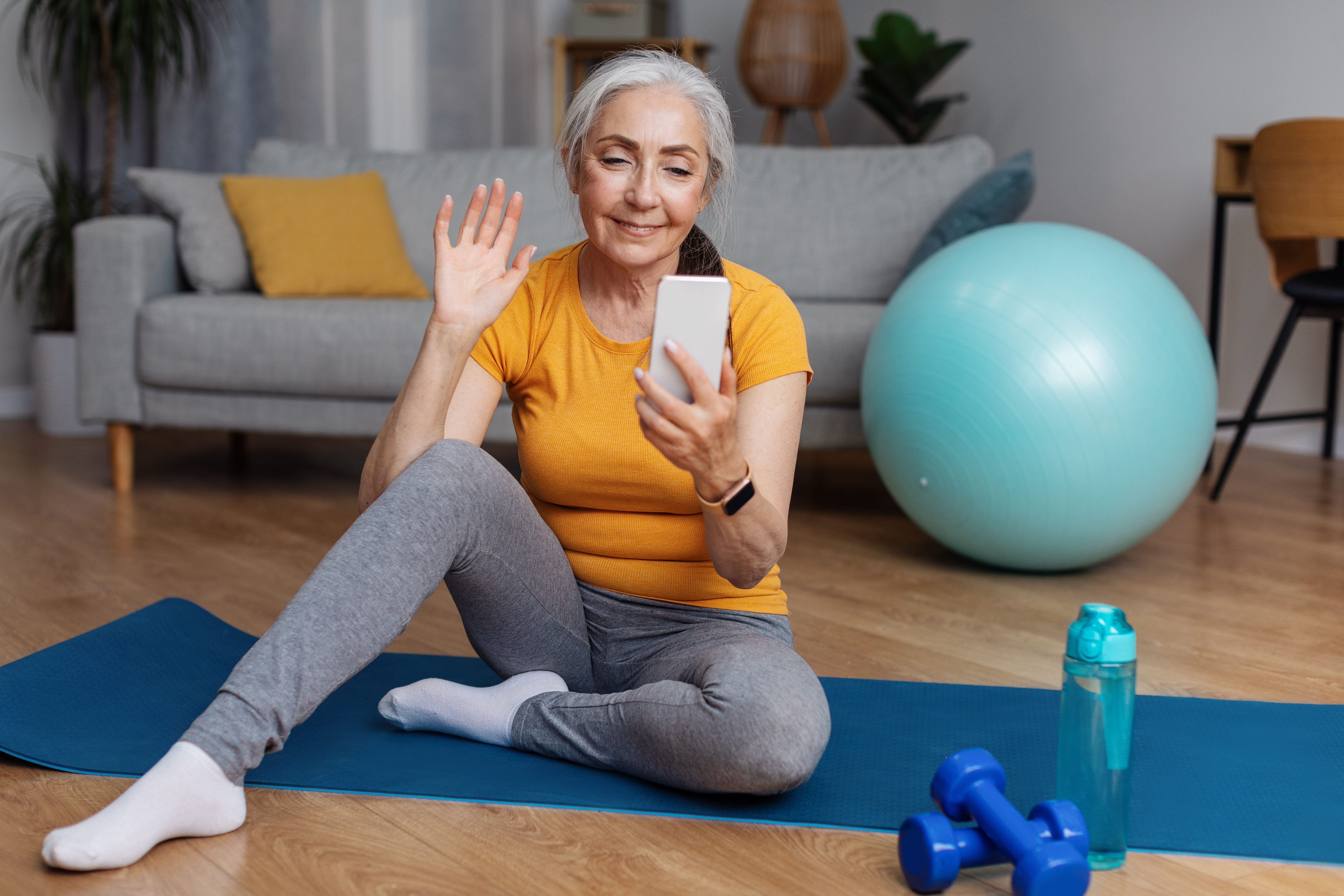 Image of a woman in yoga clothes, sitting on a yoga mat in a living room with a yoga ball, water bottle, and weights beside her, holding a phone in one hand and smiling and waving toward the phone while in conversation with her health coach.