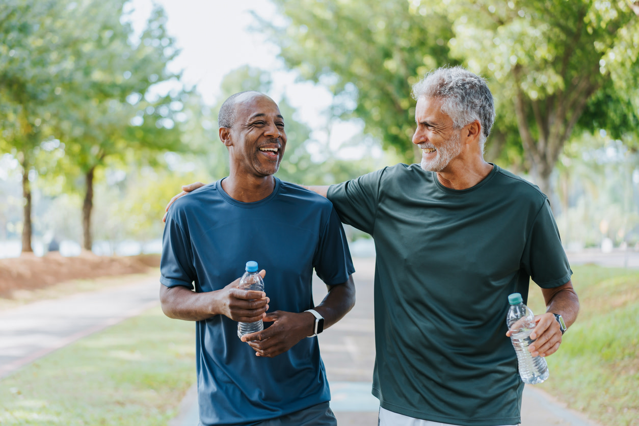 Image of two smiling men in exercise clothes walking side by side through a park, one man has his arm around the other and both are carrying a water bottle