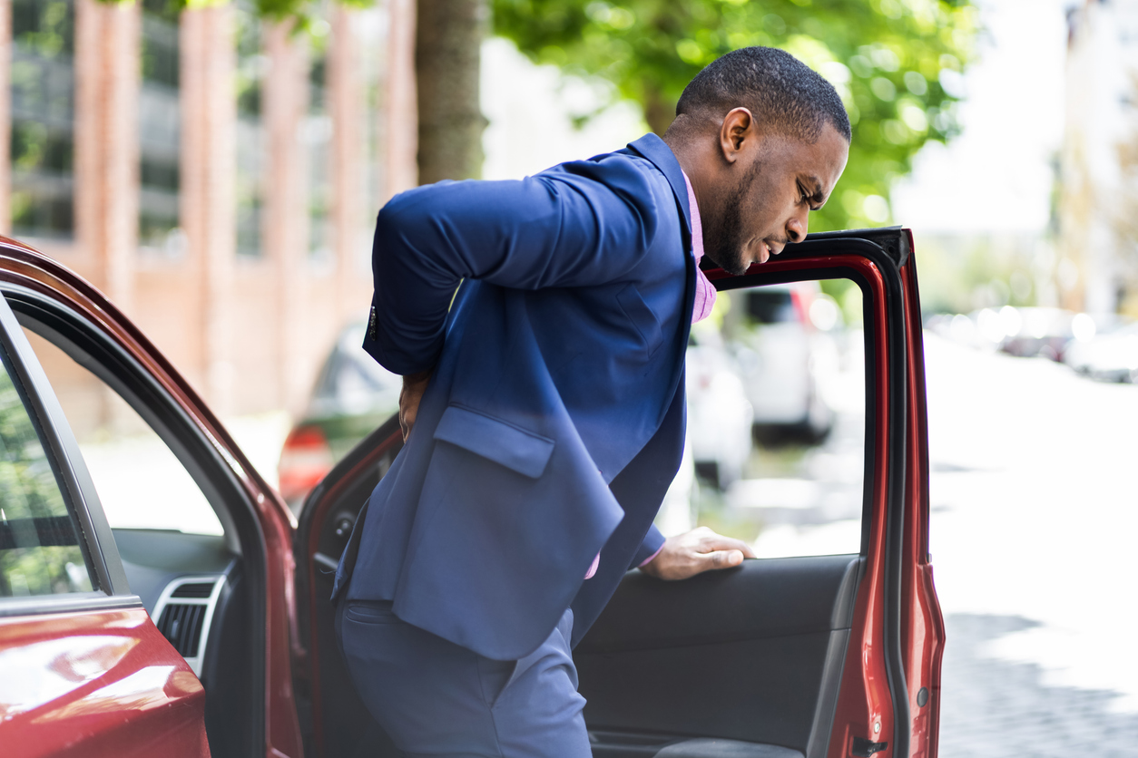 Professional man in navy blue suit entering a red car on a sunny day, with trees and buildings visible in the background