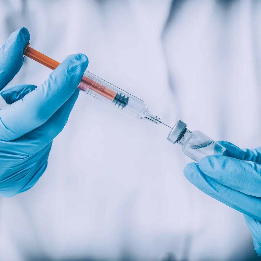Gloved hands preparing a joint injection, drawing from a glass vial against blurred white background