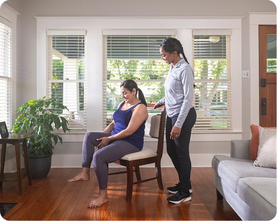 A woman is sitting in a chair in her living room while a care provider is standing behind her during a physical therapy session. 