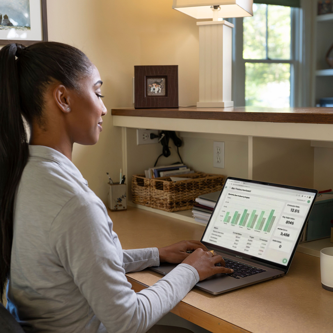 A young woman sits at a desk typing on a laptop computer. Her screen shows a business dashboard with a bar graph and various dollar amounts and percentages.