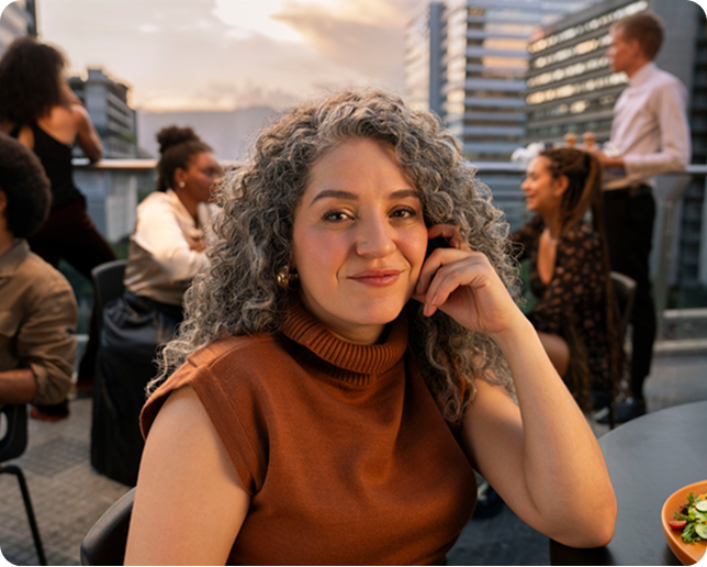 A woman with curly hair smiles at an outdoor event on an urban rooftop. Other people talk in the background.