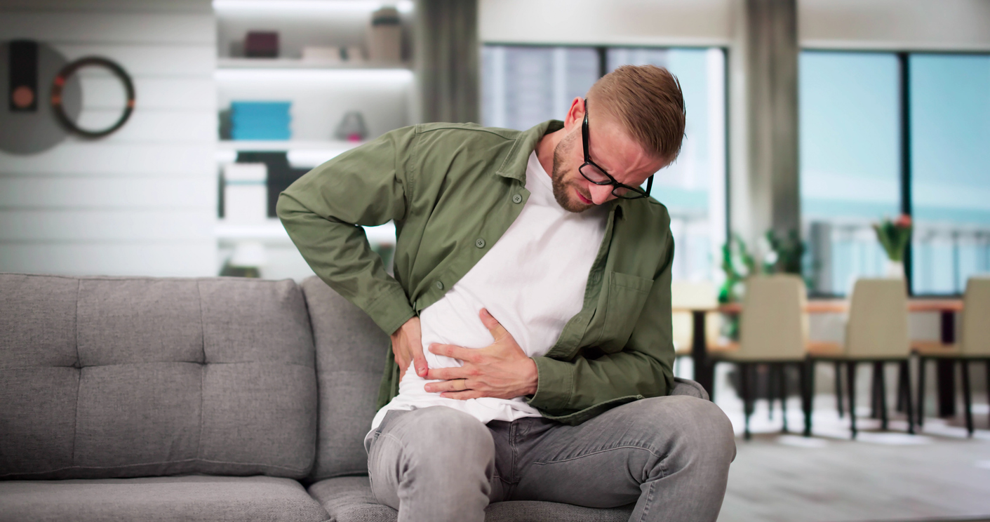 Person in green shirt and glasses sitting on gray couch, experiencing an oblique strain. 