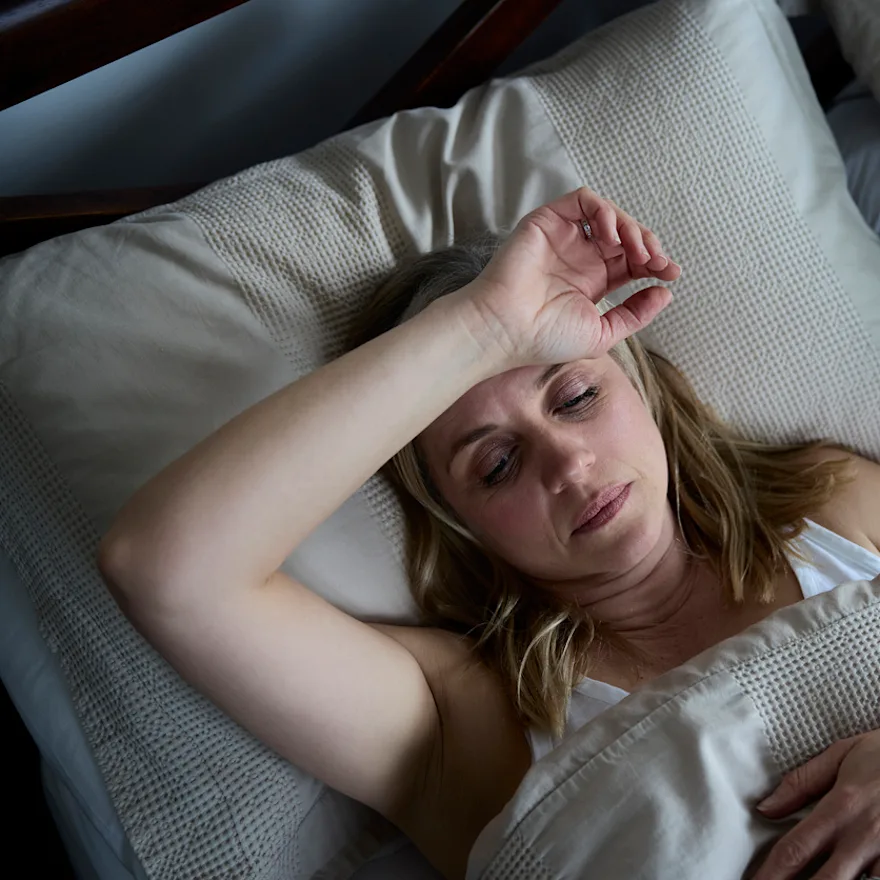Person with menopause lying in bed with hand on forehead trying to sleep, surrounded by white textured bedding